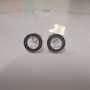 May include: A pair of silver-toned stud earrings. Each earring features a circular design with a dark border encrusted with small, sparkling stones. The earrings are set against a white background.