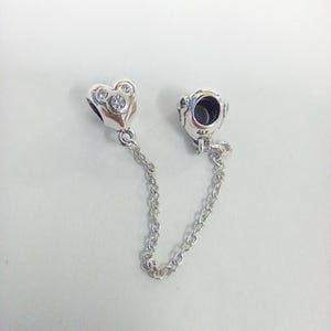 May include: A silver charm bracelet safety chain with a heart-shaped charm and a round charm. The heart charm has three clear stones. The chain is made of small, linked silver pieces.