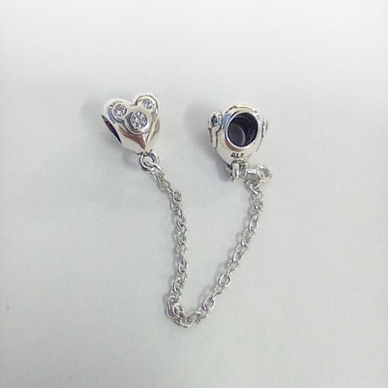 May include: A silver charm bracelet safety chain with a heart-shaped charm and a round charm. The heart charm has three clear stones. The chain is made of small, linked silver pieces.