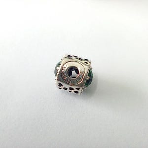 May include: A silver charm with a square design featuring cut-out hearts and the words "TIME CO." around a central circle. The charm is accented with a dark green bead.