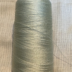 May include: A spool of pale green thread, tightly wound around a cardboard core. The thread has a subtle sheen, suggesting a silk or high-quality cotton material. The thread is unwound slightly, with a single strand extending from the spool. Suitable for sewing, embroidery, or crafting.
