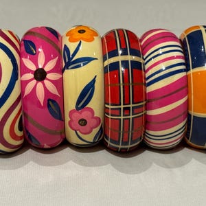 May include: Six colourful, wide bangles with various designs. The bangles feature patterns such as swirls, floral motifs, plaid, and stripes. Colours include red, pink, blue, orange, and yellow. Each bangle is approximately 7.6 cm wide.