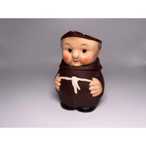 May include: A ceramic figurine of a monk in a brown robe with a white rope belt. The monk has a smiling face with rosy cheeks and a brown cowl. The figurine is standing on a white surface.
