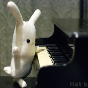 May include: A white plush bunny rabbit with black eyes sits on a black piano keyboard. The bunny is facing the right side of the image and its right paw is resting on the piano keys.