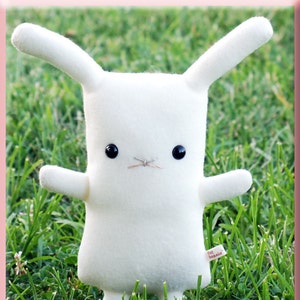 May include: White plush bunny rabbit toy with black button eyes. The bunny is standing on green grass. The toy has a small tag that says "flat bonnie".