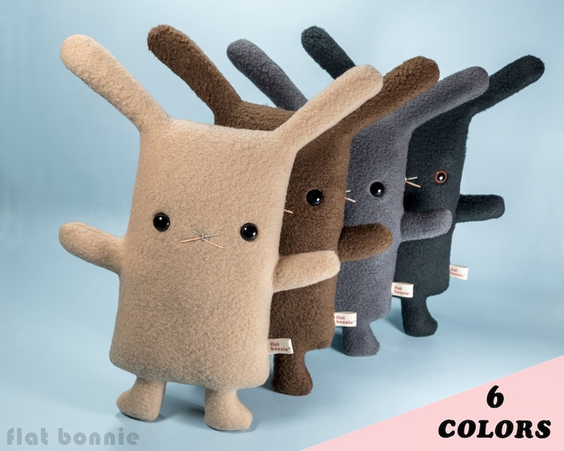 May include: Six plush bunny toys in different colors: beige, brown, gray, black, pink, and blue. Each bunny has a simple, cartoon-like face with black eyes and a stitched nose.
