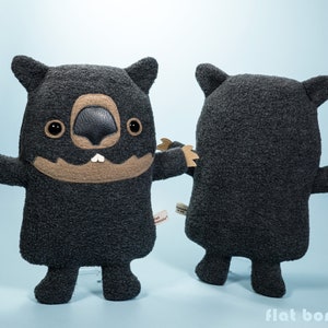 May include: A plush toy shaped like a wombat. The toy is black with a brown face and a toothy grin. The toy is standing on a light blue background.