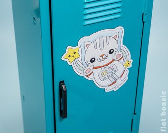 Può includere: Una porta di armadietto blu turchese con un adesivo di un gatto astronauta bianco. L'adesivo presenta un gatto bianco che indossa una tuta spaziale bianca con un cuore rosso e un pesce blu sul petto. Il gatto tiene una stella gialla con un viso sorridente. L'adesivo ha il testo "flat bonnie" nella parte inferiore.