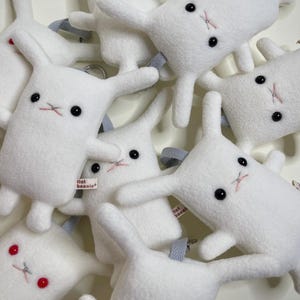 May include: A collection of white plush bunny toys with black button eyes and pink stitched mouths. Each bunny has long ears and a gray ribbon loop. Some bunnies have a small tag that reads "flat bonnie".