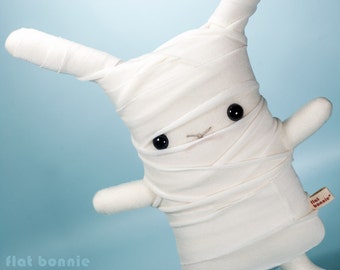 May include: White plush bunny toy wrapped in white fabric bandages, with black button eyes and a stitched nose. The toy has a tag that says "flat bonnie".