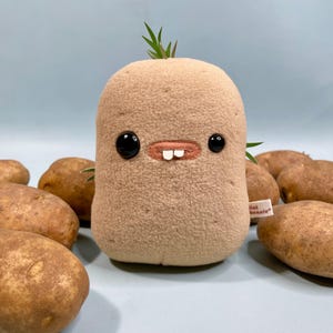 May include: A plush toy potato with black button eyes, a stitched mouth, and two white teeth. The tan-colored toy has green sprouts and is surrounded by several real potatoes on a light blue surface. A small tag reads "lil bonnie".