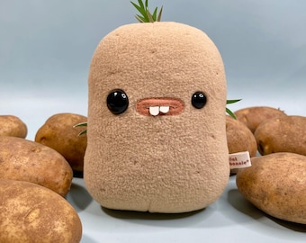 May include: A plush toy potato with black button eyes, a stitched mouth, and two white teeth. The tan-colored toy has green sprouts and is surrounded by several real potatoes on a light blue surface. A small tag reads "lil bonnie".