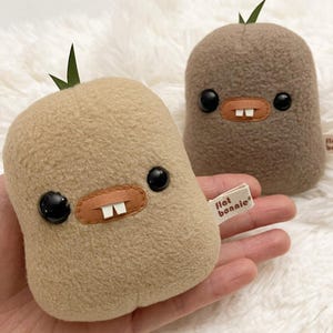May include: Two plush toys with a bean-shaped design. The toys are light brown and brown, with black button eyes, a tan mouth, and two white teeth. Each toy has a small green leaf on top. One toy has a tag that says "flat bonnie".