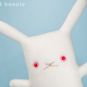 May include: White plush bunny with red button eyes and a stitched nose. The bunny is lying on its side with its head turned towards the camera.