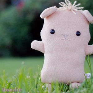 May include: A plush pink toy animal with a brown nose and black eyes. The toy is sitting in a grassy field.