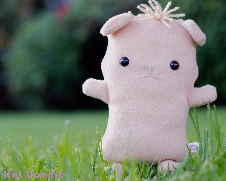 May include: A plush pink toy animal with a brown nose and black eyes. The toy is sitting in a grassy field.