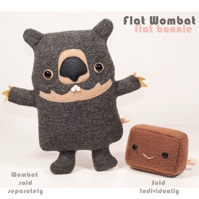 May include: A gray and brown plush toy shaped like a wombat with a flat body. The toy has a friendly face with black eyes, a brown nose, and a small mouth with two white teeth. The toy is standing on a white surface. The text "Flat Wombat Flat Bonnie" is written in pink letters above the toy. The text "Wombat sold separately" is written below the toy.