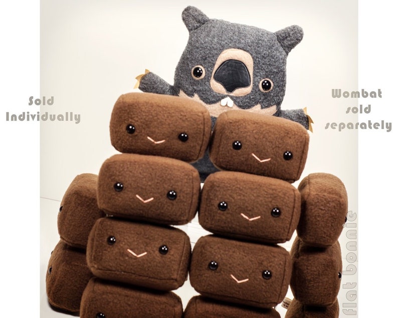 May include: A gray plush wombat toy with black eyes and a toothy grin, standing in front of a stack of brown plush blocks with smiling faces. The blocks are labeled "flat bonus" and the wombat is labeled "wombat sold separately".