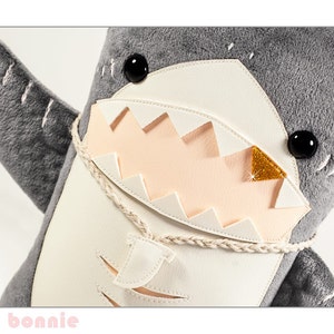 May include: A gray and white plush shark toy with a large, toothy grin. The shark has black eyes and a gold glitter accent on its mouth.