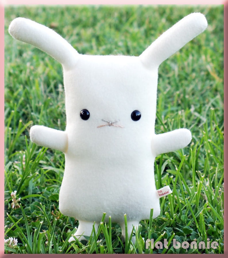 May include: White plush bunny rabbit toy with black button eyes. The bunny is standing on green grass. The toy has a small tag that says "flat bonnie".