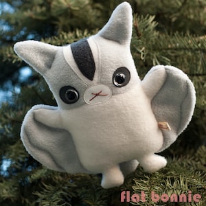 May include: A gray and white plush toy shaped like a flying squirrel with large black eyes. The toy has a black stripe down its back and a small white nose.