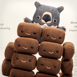 May include: A gray plush wombat toy is standing in front of a stack of brown plush blocks with smiling faces. The blocks are stacked in a pyramid shape. The text "Sold Individually" is on the left side of the image and the text "Wombat sold separately" is on the right side of the image.