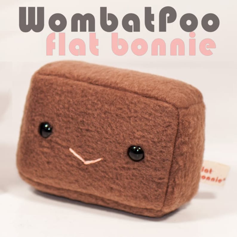 May include: A brown plush toy shaped like a rectangular block with a smiling face. The toy has black eyes and a pink stitched smile. The toy is labeled "Wombat Poo Flat Bonnie".