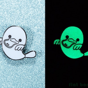 Puede incluir: Un pin de esmalte blanco de un ornitorrinco con un contorno negro. El ornitorrinco está en una pose de fantasma con los brazos levantados. El pin brilla en verde en la oscuridad.
