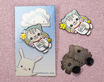 Puede incluir: Dos pines de esmalte con un gato astronauta de dibujos animados con un corazón y un símbolo de pez en el pecho. El gato lleva un traje espacial azul y blanco y está rodeado de estrellas. Los pines están sobre un fondo rosa con una tarjeta blanca detrás de uno de los pines. La tarjeta tiene una nube y un conejo y el texto "flatbonnie.com".