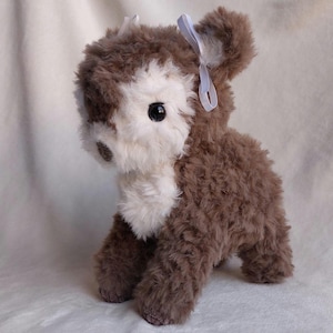 May include: A soft toy deer with brown and white fur. The deer has large black eyes and a small white bow on its ear. The toy is positioned on a white surface.