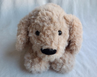 Puede incluir: Un perro de peluche con suave pelaje marrón claro. El perro tiene ojos negros de botón y una nariz negra. El juguete está sobre una superficie blanca y texturizada. El pelaje del perro parece esponjoso y tierno.