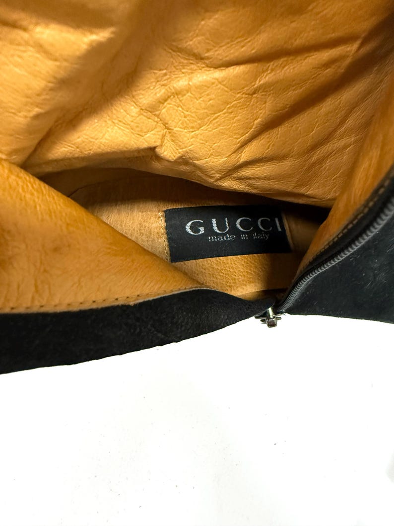 Może przedstawiać: Wnętrze czarnego sk&oacute;rzanego buta z beżową podszewką. Etykieta Gucci jest widoczna wewnątrz buta. But ma zapięcie na zamek błyskawiczny.