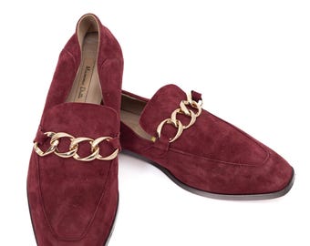 May include: A pair of burgundy suede loafers with gold-tone chain detailing. The shoes have a classic slip-on design with a rounded toe and a low heel. The brand name "Massimo Dutti" is visible inside the shoe.