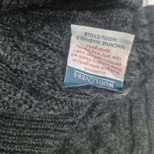 May include: Close-up of a dark gray knit wool garment. A white tag with black text is attached, with the words "MACHINE WASHABLE WOOL CYCLE" and a warning about detergents. The tag also has the brand name "WOOLOVERS".