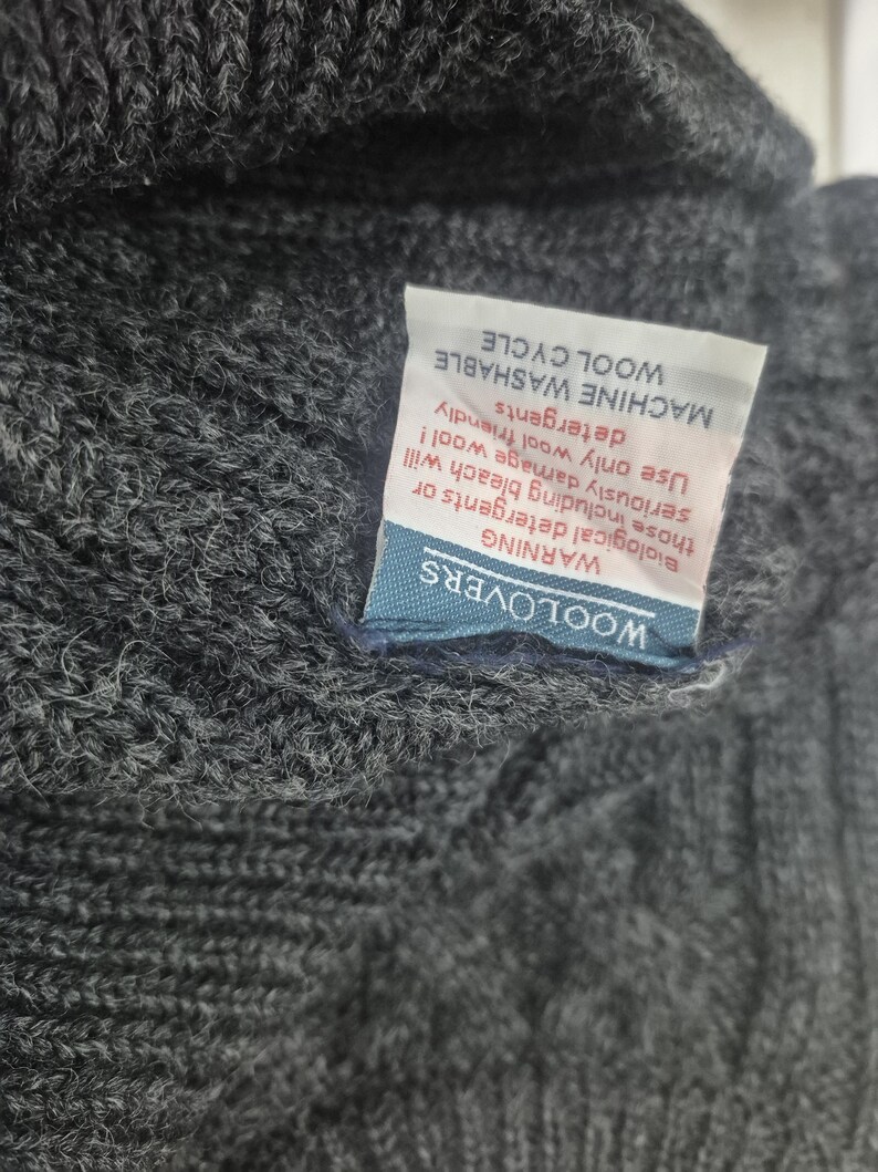 May include: Close-up of a dark gray knit wool garment. A white tag with black text is attached, with the words "MACHINE WASHABLE WOOL CYCLE" and a warning about detergents. The tag also has the brand name "WOOLOVERS".
