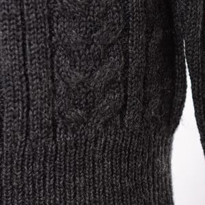 May include: Close-up of a dark gray cable-knit sweater. The textured knit features a ribbed hem and a detailed cable pattern on the front. The sleeves are visible on the right side of the frame. The sweater appears to be made of a soft, warm material.