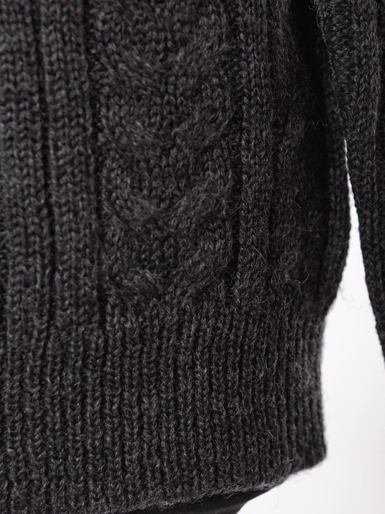 May include: Close-up of a dark gray cable-knit sweater. The textured knit features a ribbed hem and a detailed cable pattern on the front. The sleeves are visible on the right side of the frame. The sweater appears to be made of a soft, warm material.