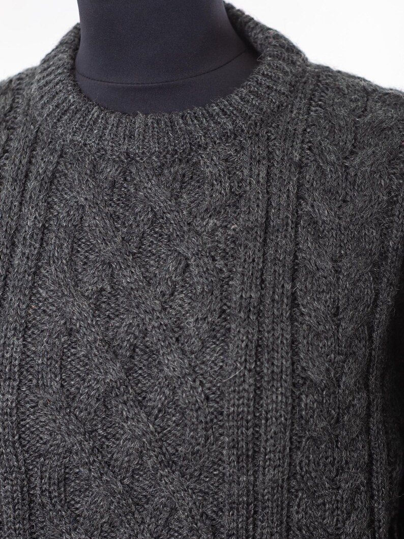 May include: A close-up of a charcoal gray cable-knit sweater. The sweater features a crew neck and a textured pattern of vertical and diamond-shaped cable designs. The knitwear is displayed on a mannequin.