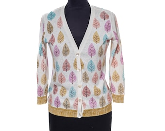 May include: A white V-neck cardigan featuring a repeating pattern of stylized leaves in shades of blue, pink, and gold. The garment has long sleeves, a button-down front with light-colored buttons, and a textured gold-toned hem and cuffs.