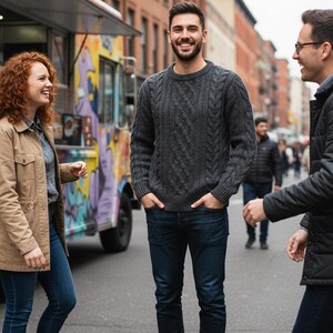 May include: A group of people standing on a city street. A man in a gray cable-knit sweater and dark blue jeans smiles. Others wear jackets and jeans. A colorful food truck is in the background.