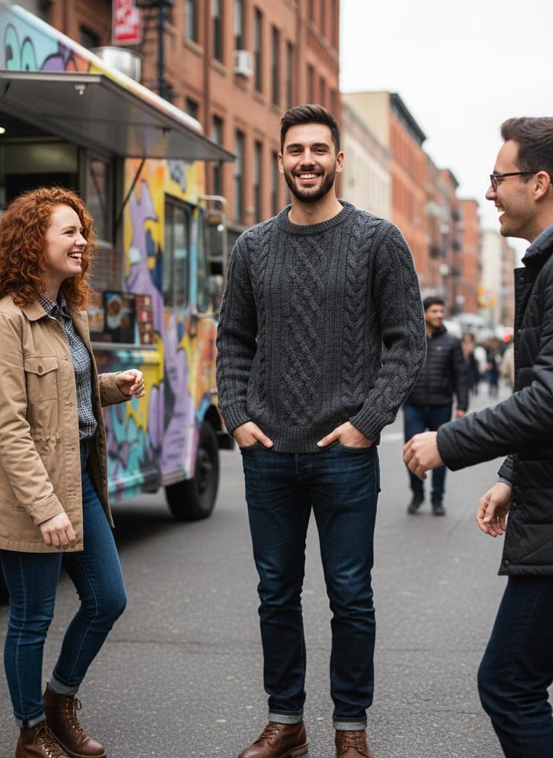May include: A group of people standing on a city street. A man in a gray cable-knit sweater and dark blue jeans smiles. Others wear jackets and jeans. A colorful food truck is in the background.
