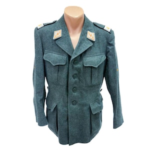 May include: A teal-coloured vintage military jacket with a notched collar, four front pockets, and button closures. The jacket has shoulder epaulets with insignia and is displayed on a mannequin. A classic piece of military apparel, it is a historical garment.