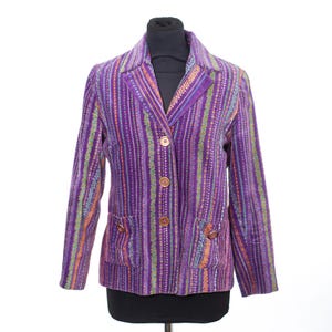 May include: A purple blazer featuring a vertical striped pattern in shades of orange, green, and light purple. The garment includes a notched lapel, three brown buttons down the front closure, and two front pockets with buttoned flaps on each side.