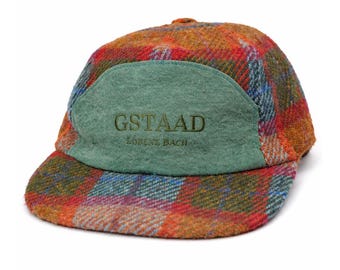 May include: A plaid baseball cap in shades of orange, red, blue, and green. The front panel is a solid green with the word "GSTAAD" embroidered in a darker green, with "LORENZ BACH" below it. The cap has a curved brim.