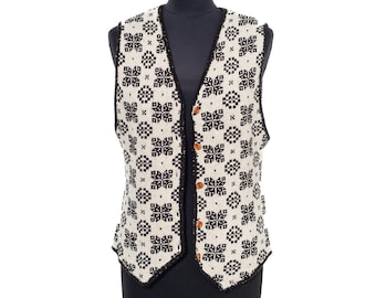 May include: A sleeveless knit vest featuring a repeating black and white geometric floral pattern. The garment has a V-neckline, a black trim along the edges, and five brown buttons down the front center. The vest is displayed on a mannequin.