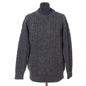 May include: A gray cable-knit sweater with a crew neck. The sweater is displayed on a mannequin and features a textured pattern across the front and sleeves. The sleeves are long and the overall style is classic.