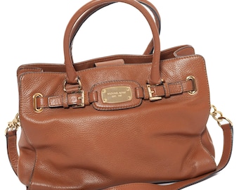 May include: A brown leather handbag with two top handles and a detachable shoulder strap. The bag features gold-tone hardware, including buckles and a chain detail. A rectangular gold plaque with the brand name is centered on the front.