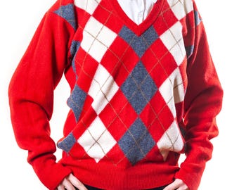 May include: A red argyle sweater with a V-neck design. The sweater features a pattern of red, white, and blue diamonds. A white collared shirt is visible underneath the sweater. The sleeves are long and the sweater is made of a soft material.