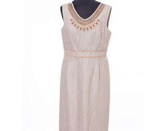 May include: A sleeveless, knee-length dress in a light beige color. The dress features a V-neckline adorned with a beaded design in gold and brown tones. A matching beaded band accents the waist. The dress is displayed on a black mannequin.