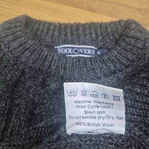 May include: A dark gray wool sweater with a crew neck. The label reads "WOOLOVERS". A tag indicates the sweater is machine washable at 40 degrees Celsius and made of 100% British wool.